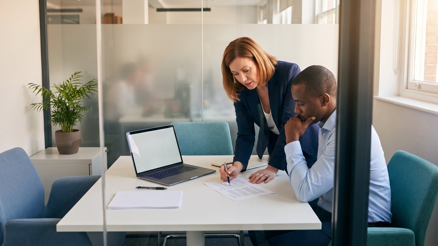 Two colleagues in a glass-walled meeting room review a printed document together before signing it off